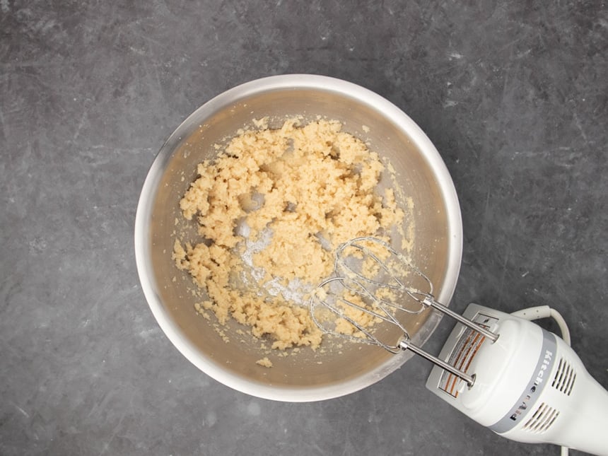 white handheld mixer with light brown butter and brown sugar beaten into choppy light brown mixture in large round mixing bowl